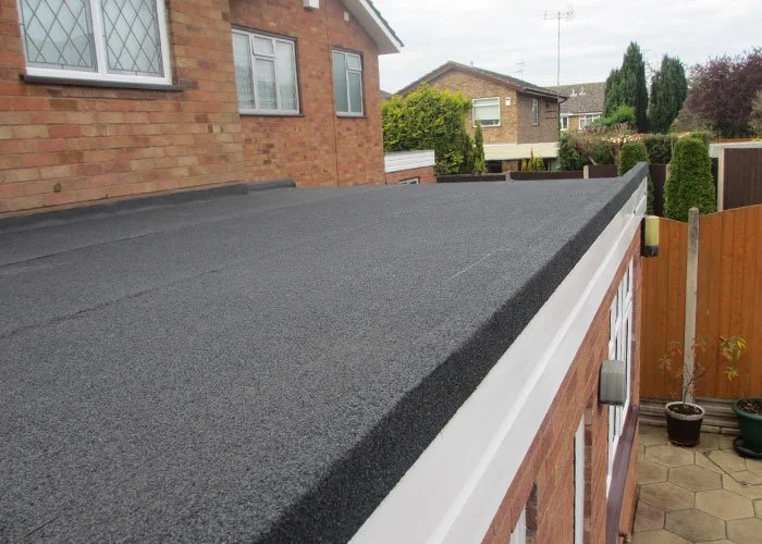 Flat roof with black roofing material on a brick house, with neighboring houses and trees in the background.