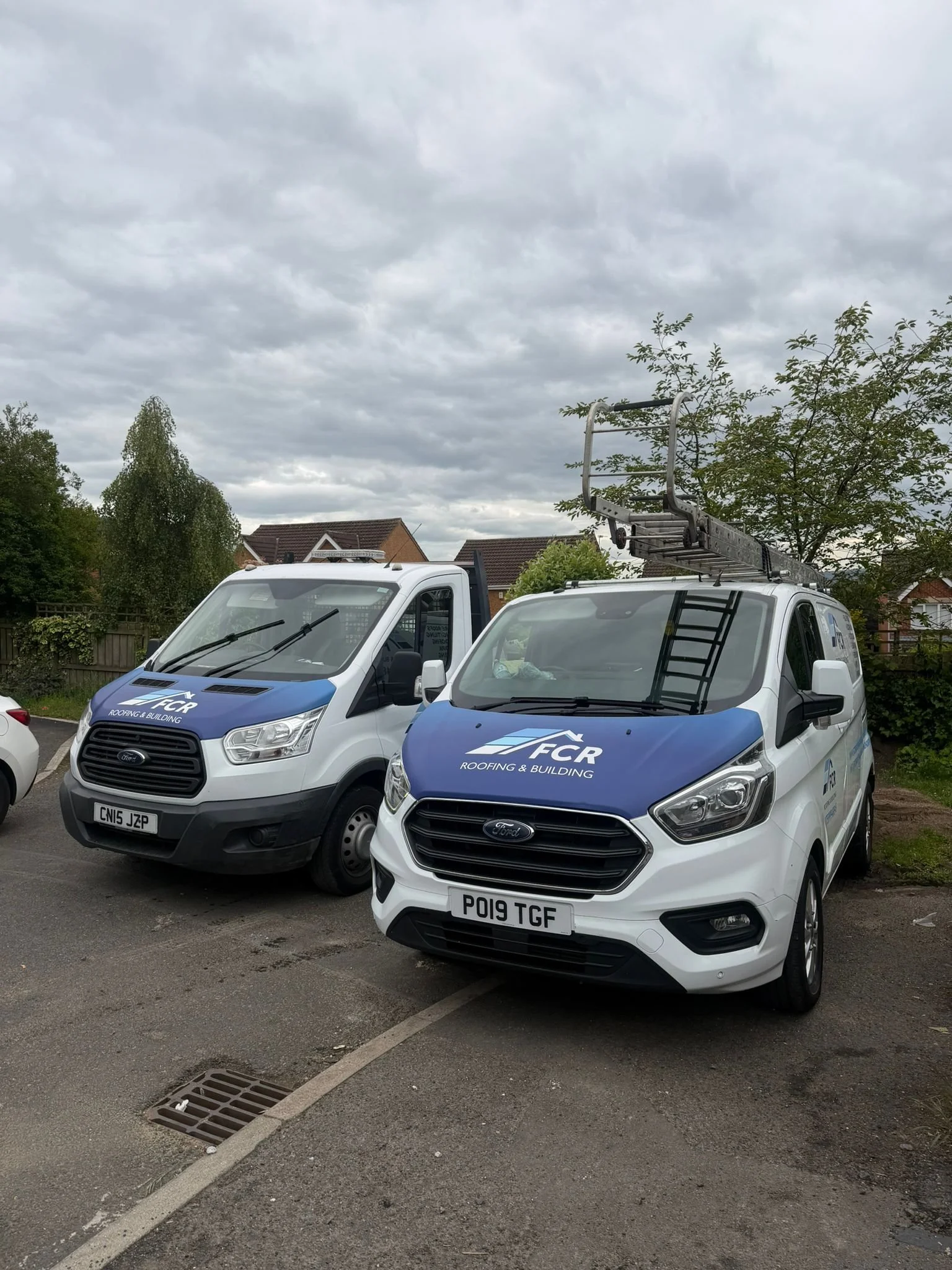 Two white utility vans with blue branding parked in a parking lot, with a ladder mounted on the roof of one van, and cloudy sky overhead, surrounded by trees and residential houses.