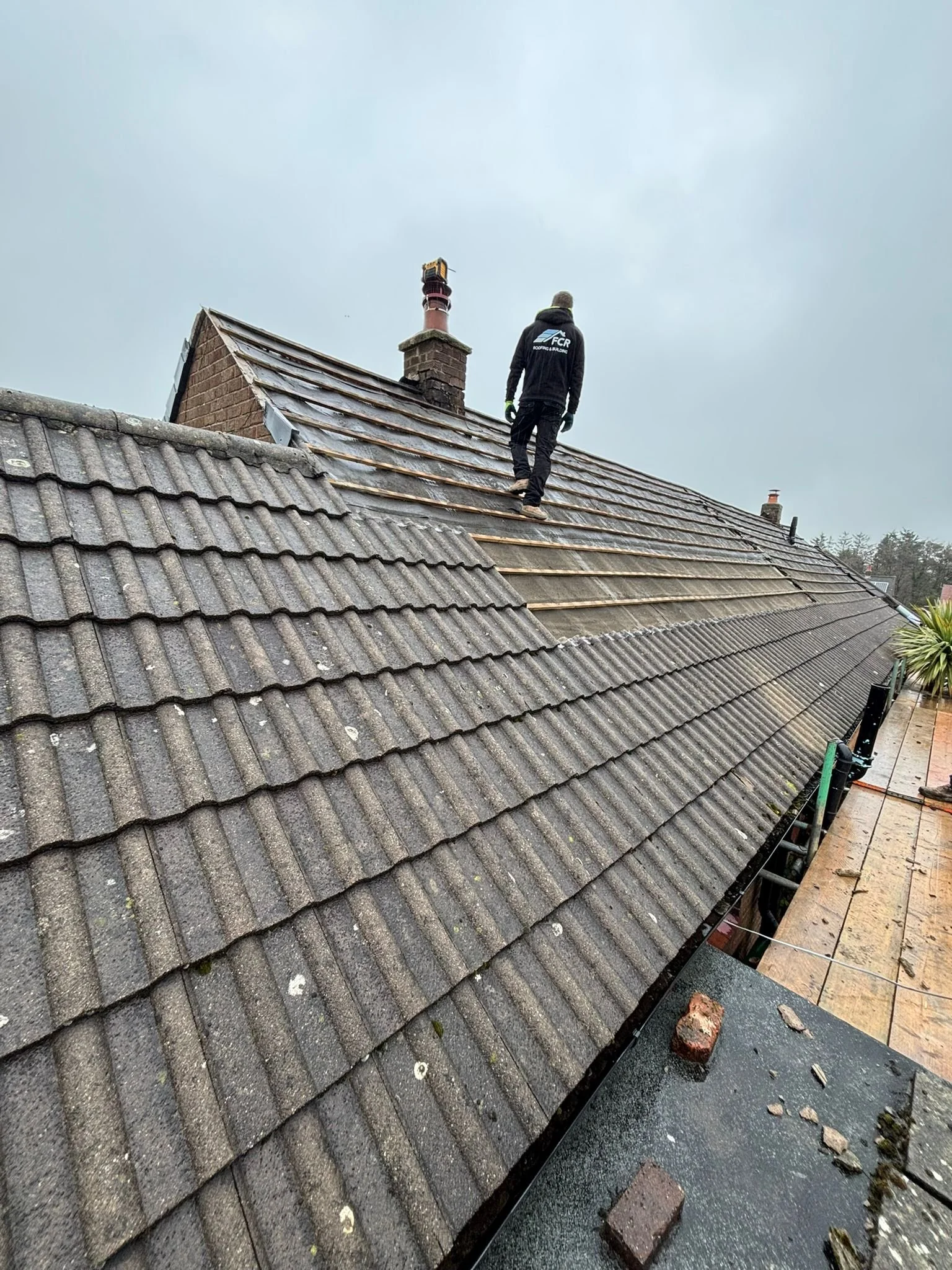 A worker on a roof replacing or repairing roof tiles. The worker is wearing a black hooded jacket, gloves, and pants, with his back to the camera. The roof has dark gray tiles, with some removed or loose tiles and scattered bricks and debris around.