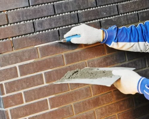 Person in blue gloves applying mortar to a brick wall with a trowel.