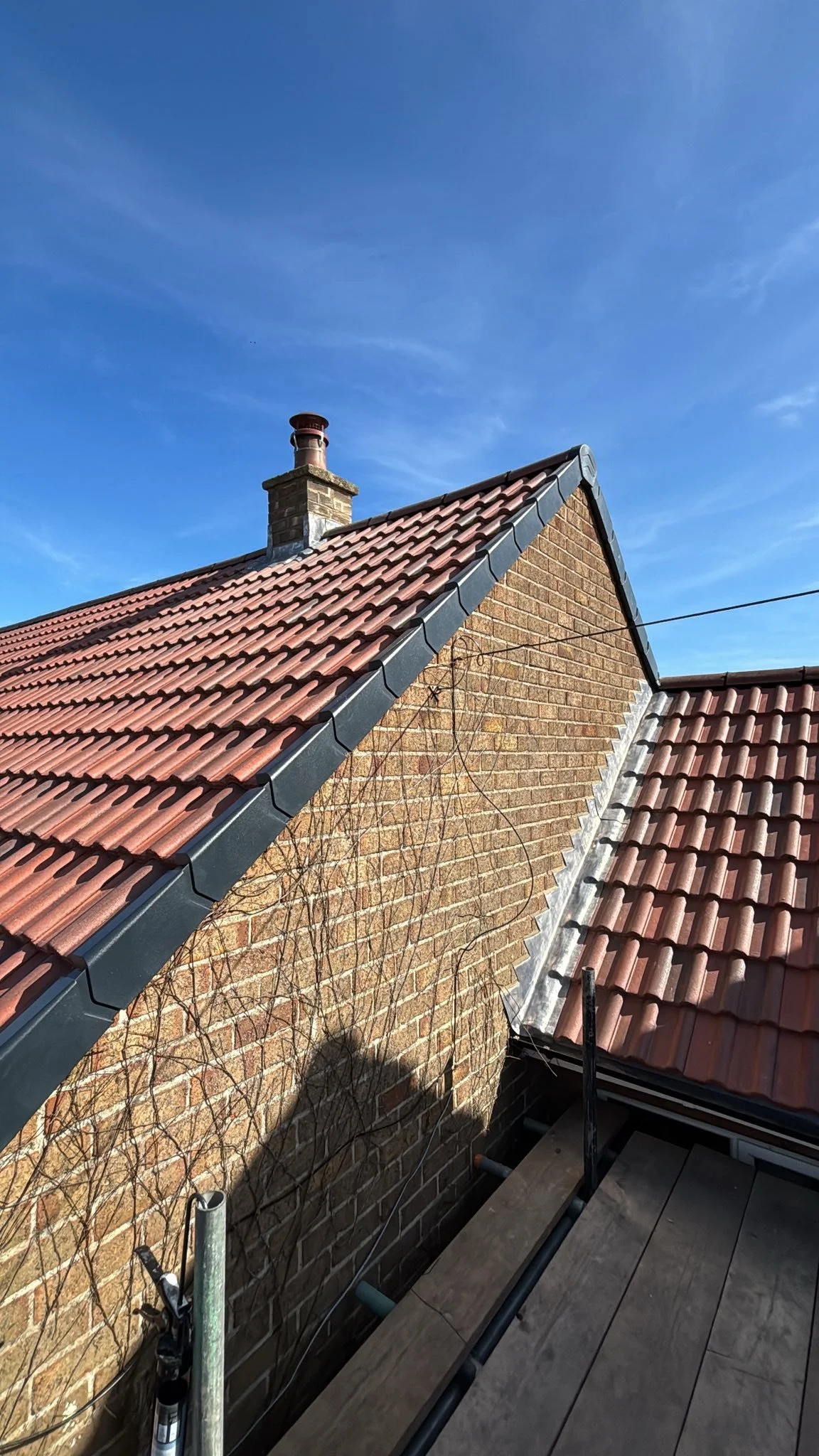 Photograph of a house roof with red clay tiles, a brick chimney, and a blue sky in the background.