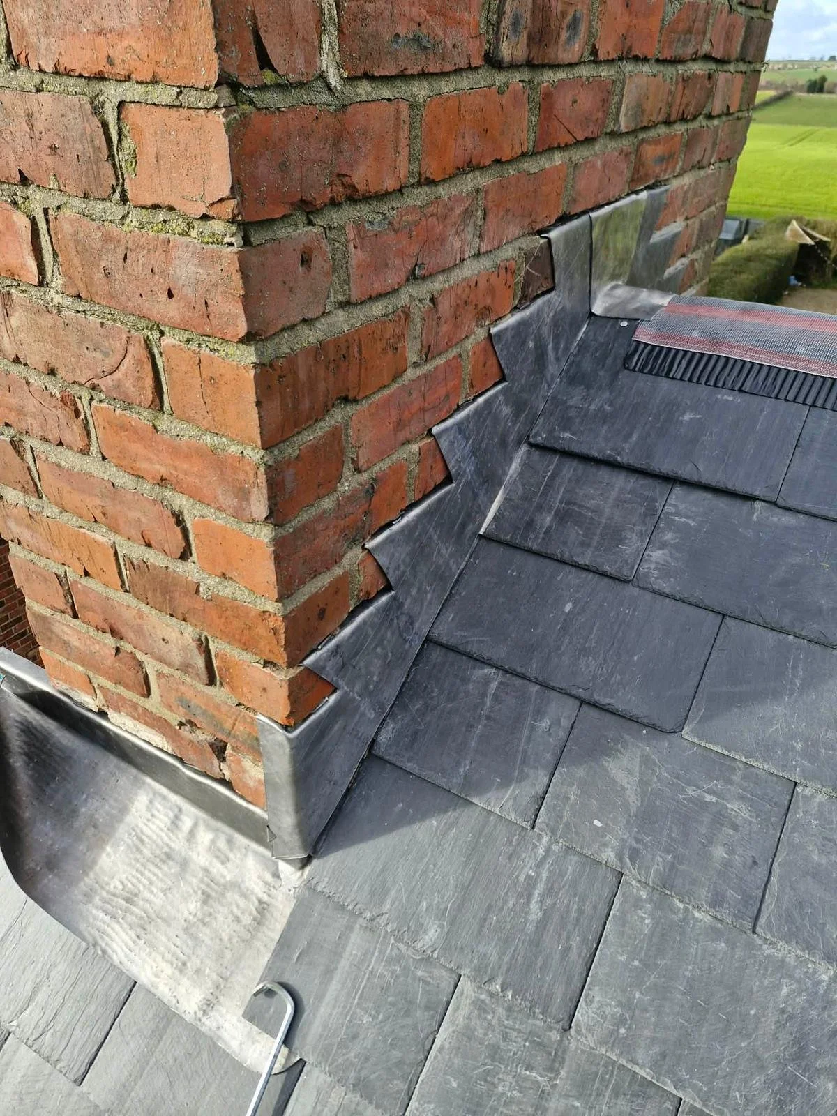 Close-up of a brick chimney with metal flashing and slate roof shingles. Green fields and sky are visible in the background.