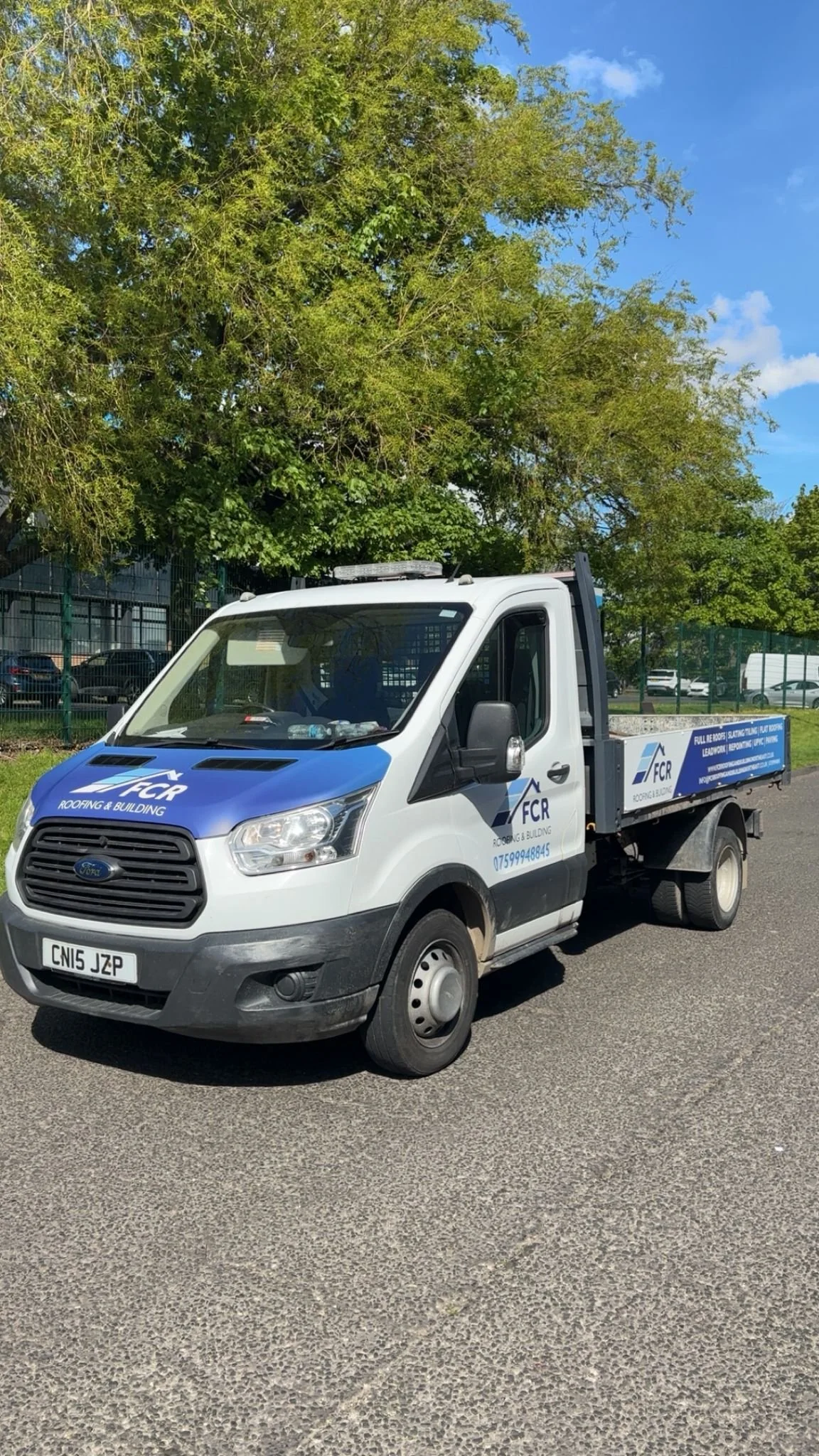 A white flatbed truck with blue branding for FCR Roofing & Building parked on a paved area, with a large leafy tree and a fence in the background under a partly cloudy blue sky.