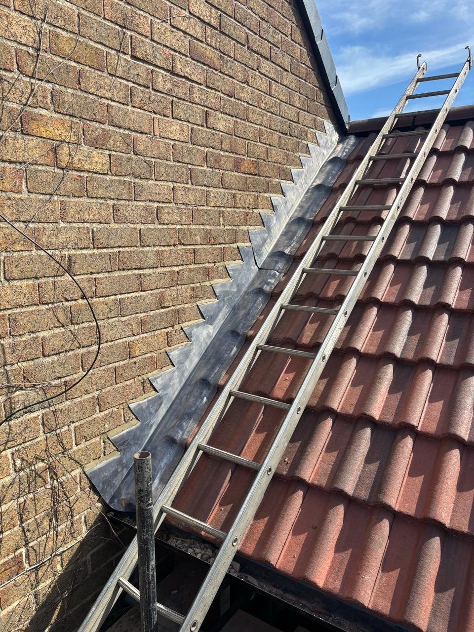 A metal ladder leaning against a brick house with a sloped tiled roof, with flashing installed along the roof edge for waterproofing.
