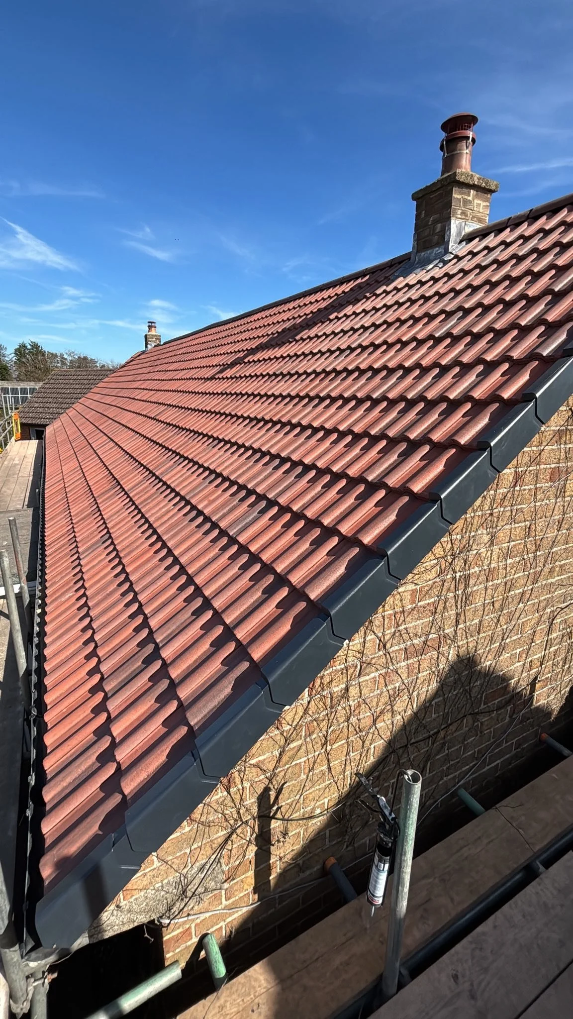 Close-up view of a red tiled sloped roof with a black metal gutter on the side, a brick chimney with a cap, and a blue sky in the background.