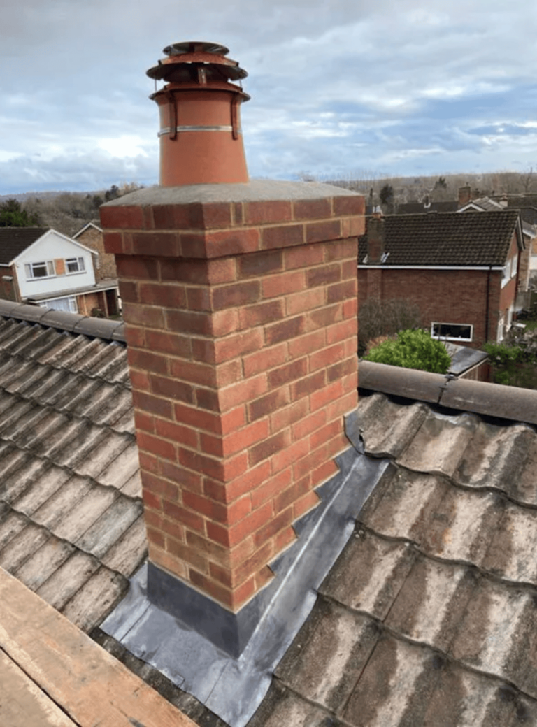 Brick chimney on a residential roof with clay tiles, metal flashing, and a cloudy sky in the background.
