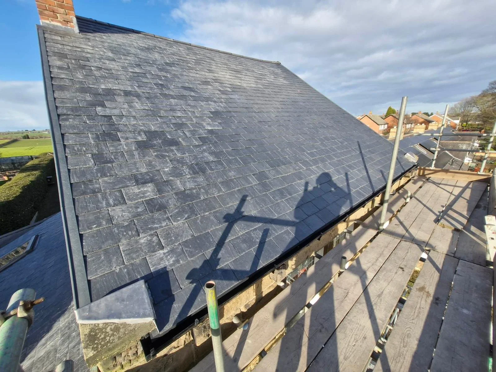A house roof under construction with slate tiles, scaffolding, and construction equipment, with neighboring houses and trees in the background under a partly cloudy sky.