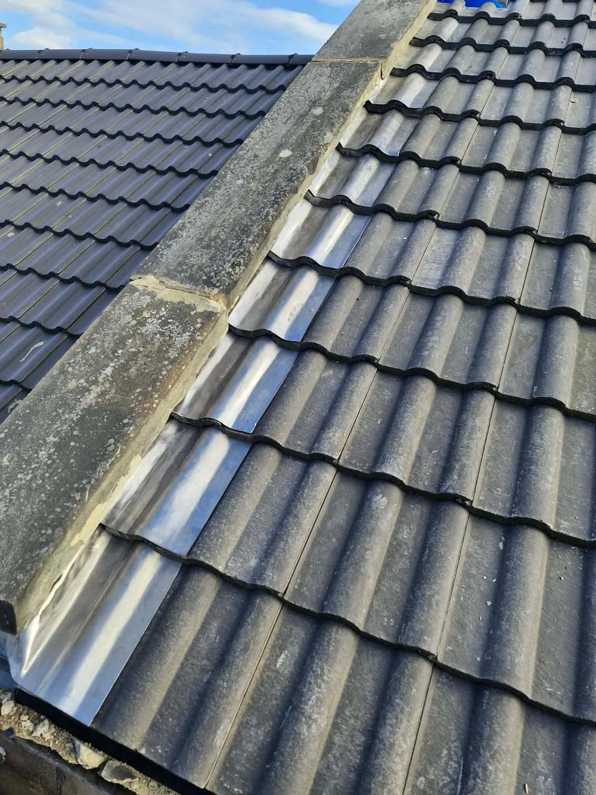 Close-up of a roof with weathered concrete tiles and a central stone chimney with moss and lichen growth, under a partly cloudy sky.