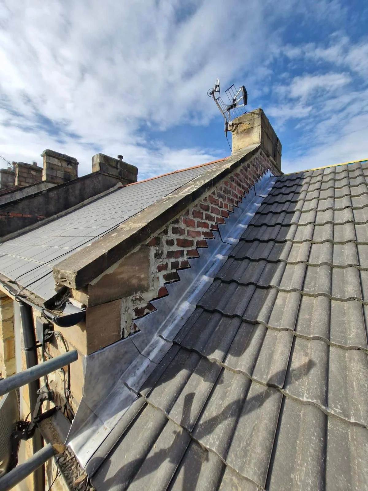 Photo of a roof with a chimney, solar panel, and antenna against a blue sky with clouds.