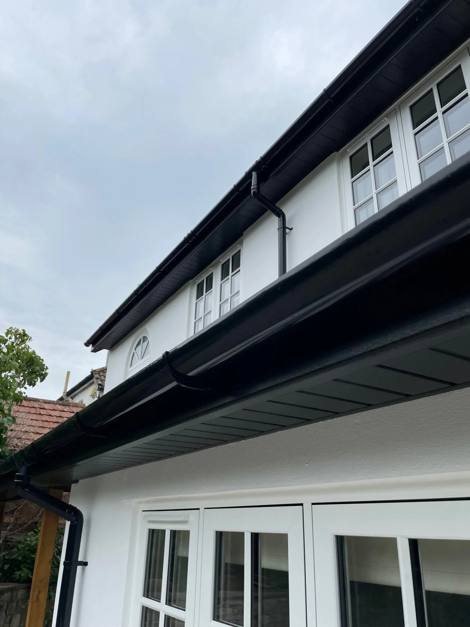Close-up of the side of a house showing a large window with multiple panes, black rain gutters, and a section of the roof, with cloudy sky in the background.