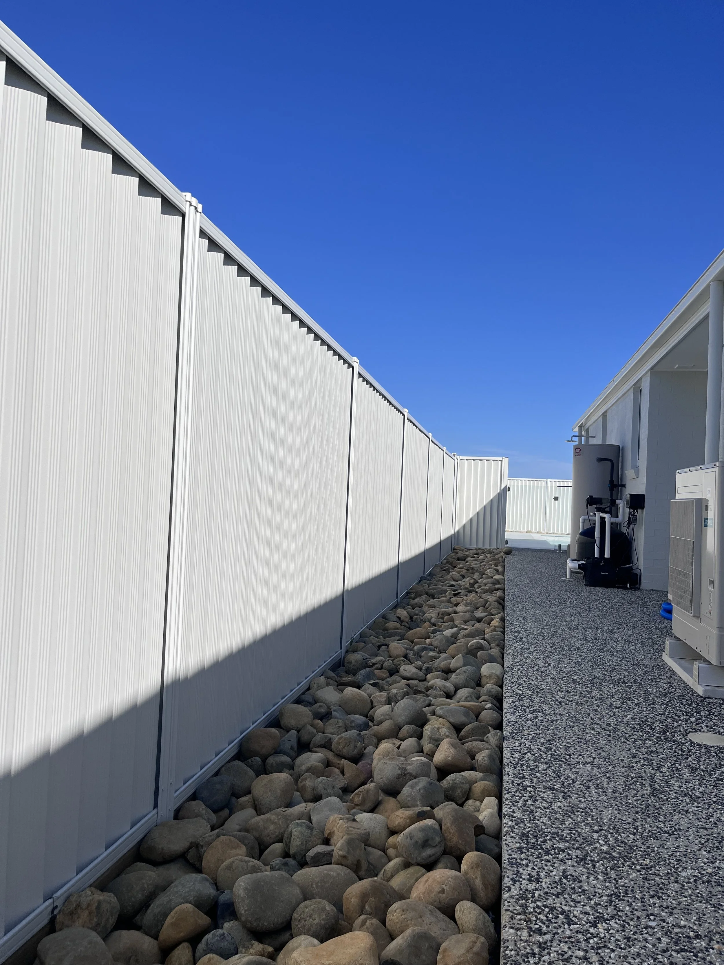 Side yard with white fence, rocks, and a house with HVAC equipment in a sunny day.