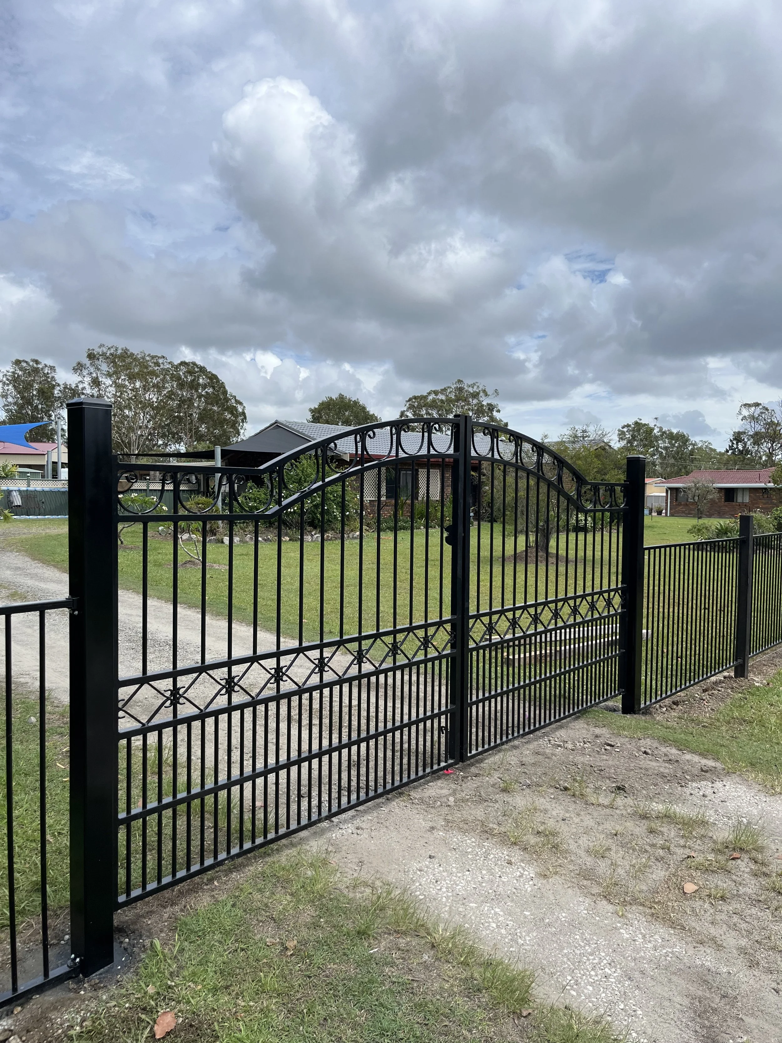 Black wrought iron gate in front of a yard with grass and gravel, some trees, houses, and a cloudy sky in the background.