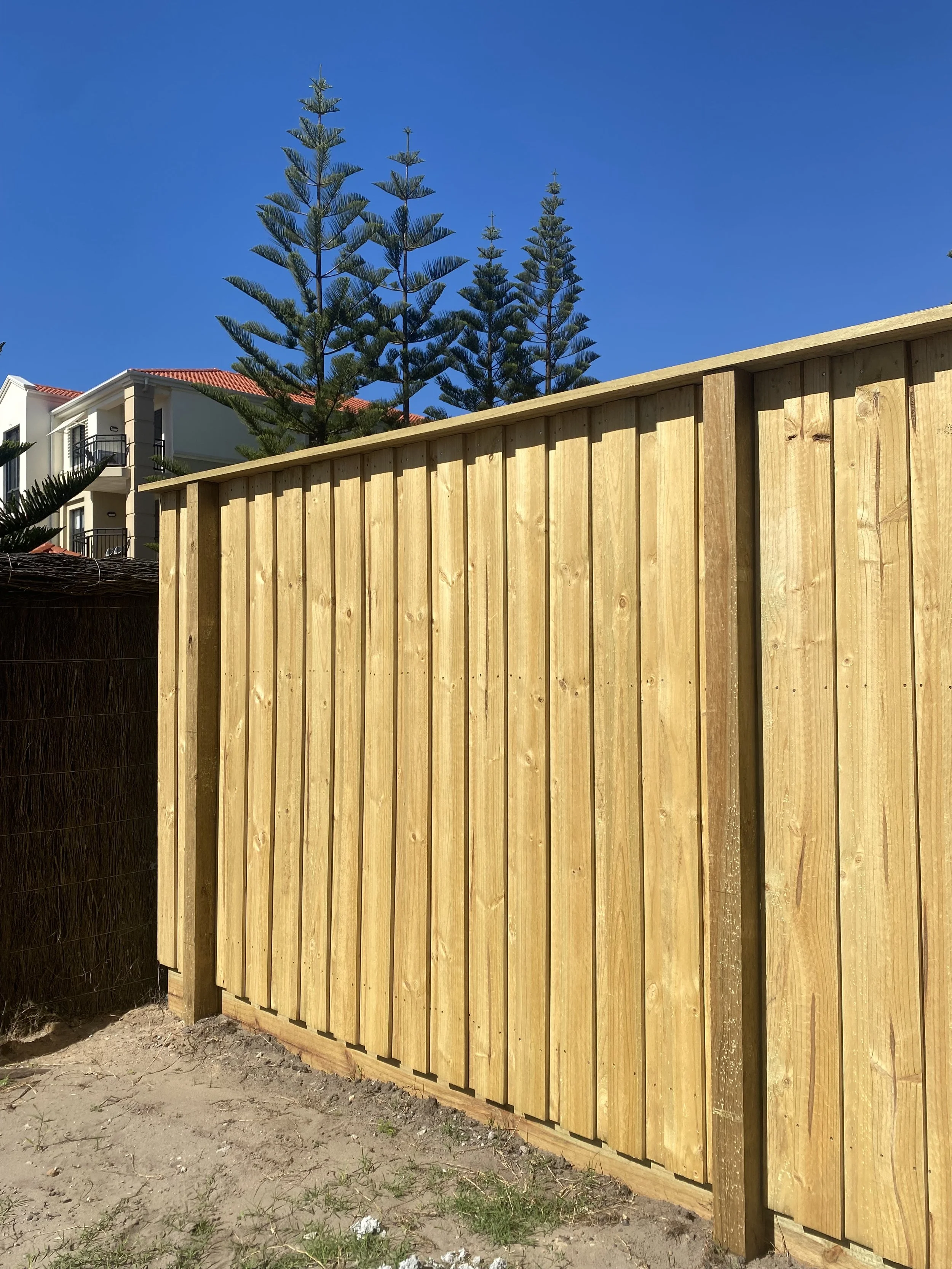 A wooden fence made of vertical boards with a curved top border in front of a modern residential building with white walls and a red-tiled roof. Tall pine trees are visible in the background under a clear blue sky.