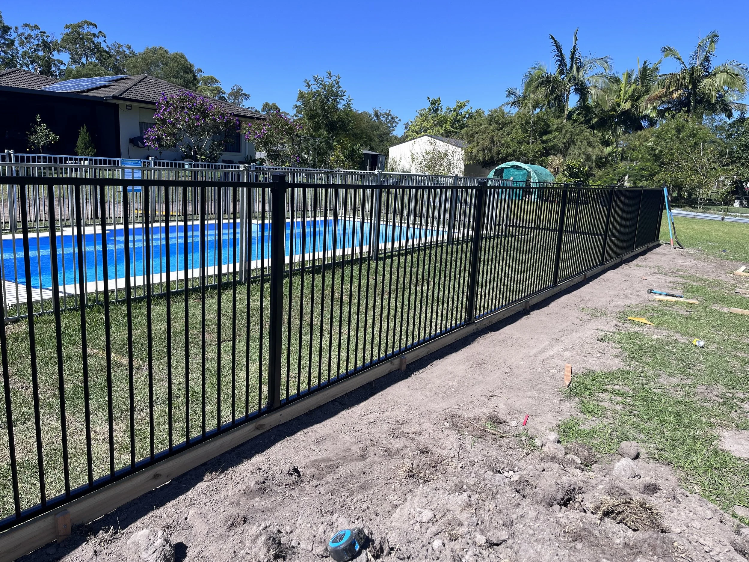 A backyard with an in-ground swimming pool surrounded by a black metal fence. There are trees and houses in the background, and a blue sky overhead.