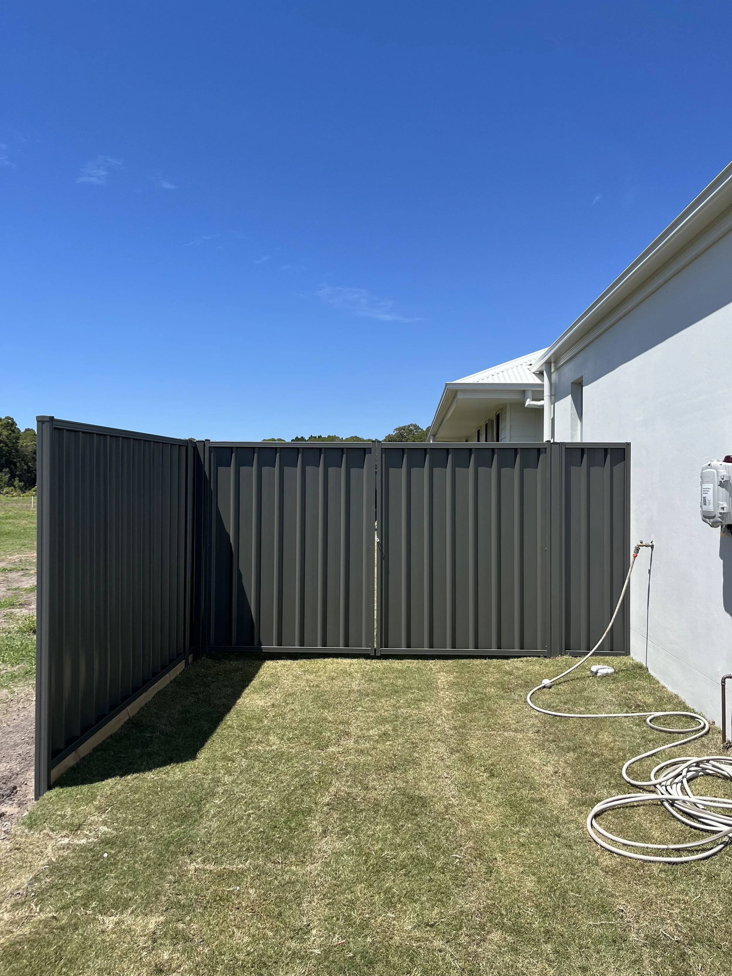 A backyard with a black metal fence and an adjacent white house. There is a garden hose on the grass and the sky is clear and blue.