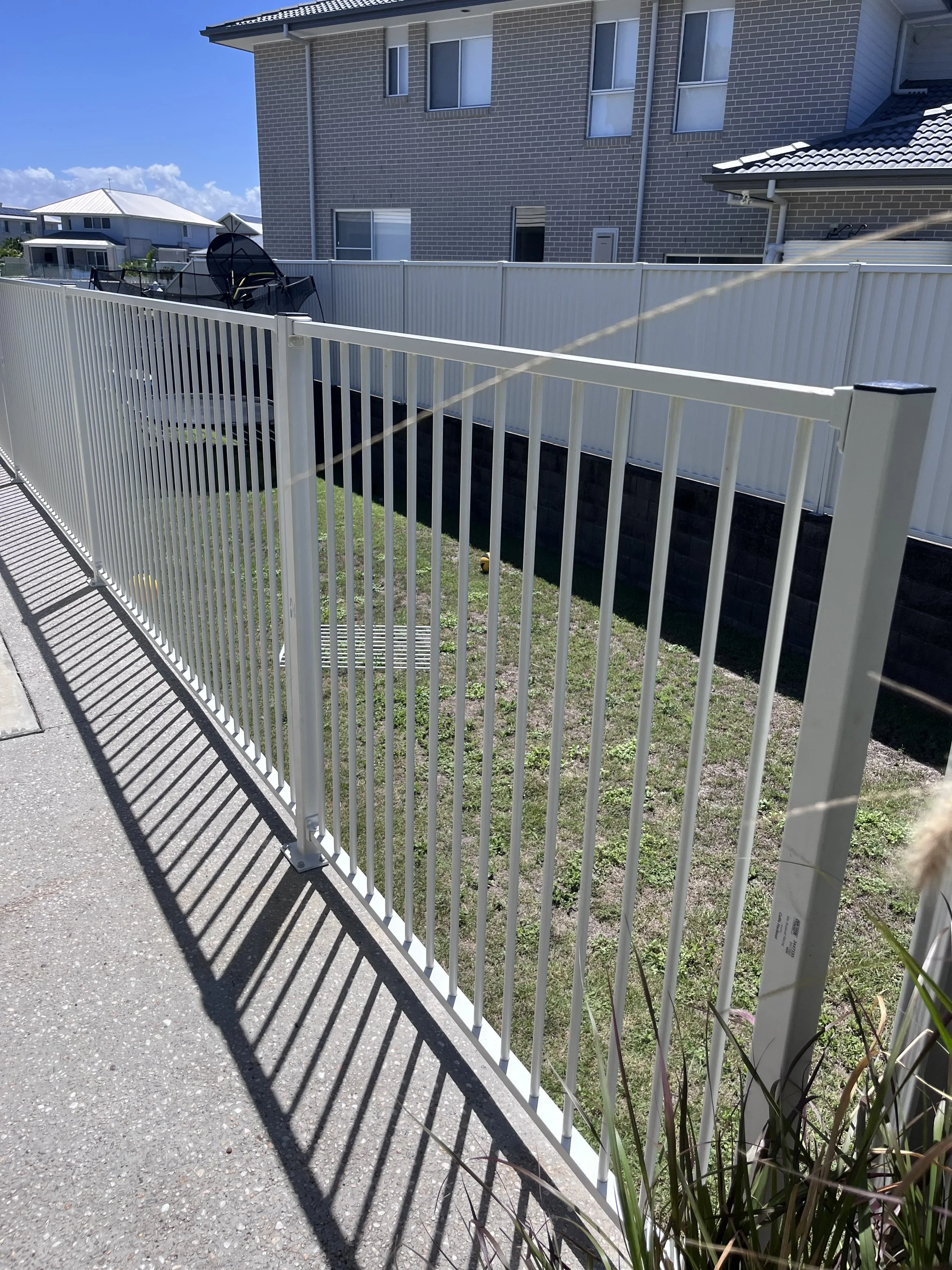 A white metal fence adjacent to a concrete sidewalk. Behind the fence, a small grassy area with some plants. In the background, a multi-story brick residential building with windows and a tiled roof. Clear blue sky with some clouds.