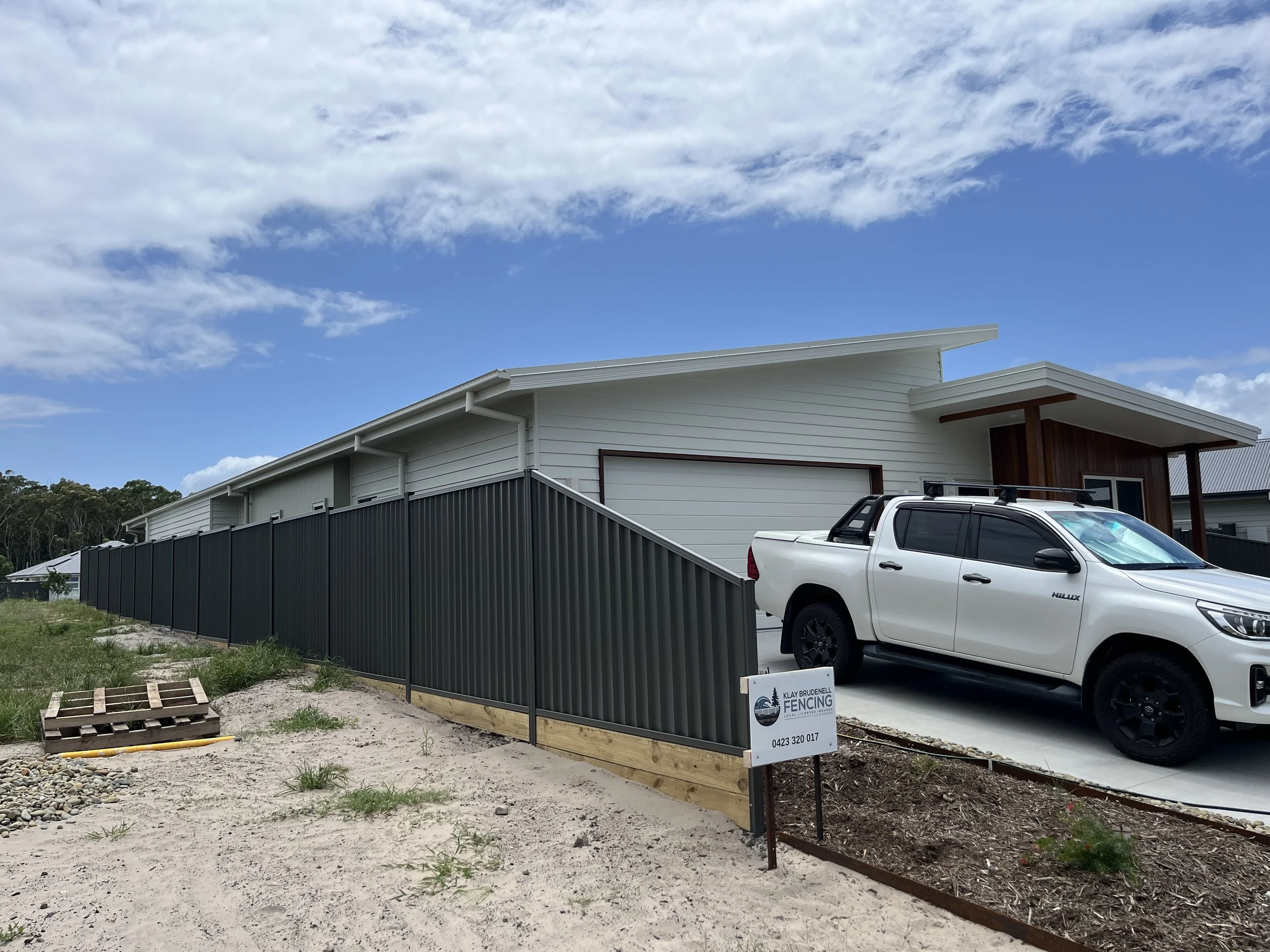 Modern house with a white exterior, a flat roof, and a grey driveway. A white pickup truck is parked in front. There is a black fence and a construction sign that reads 'Klay Brudenell Fencing.' The sky is partly cloudy with blue sky visible.