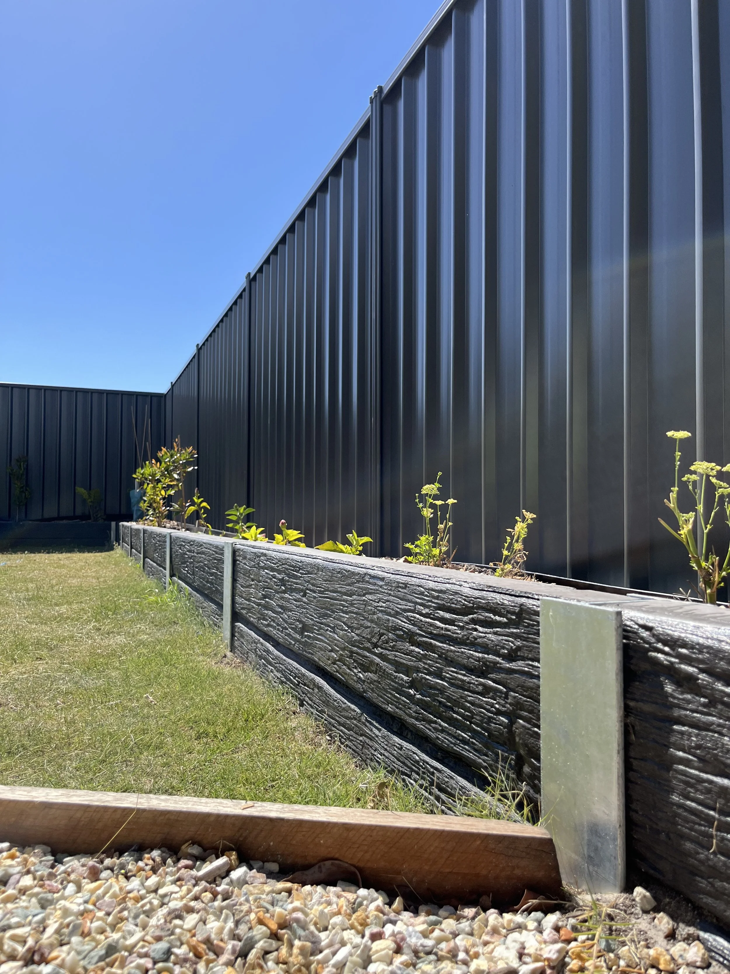 Close-up view of a black metal fence with vertical panels, a section of a wooden garden bed filled with small plants, and a gravel pathway in the foreground under a clear blue sky.