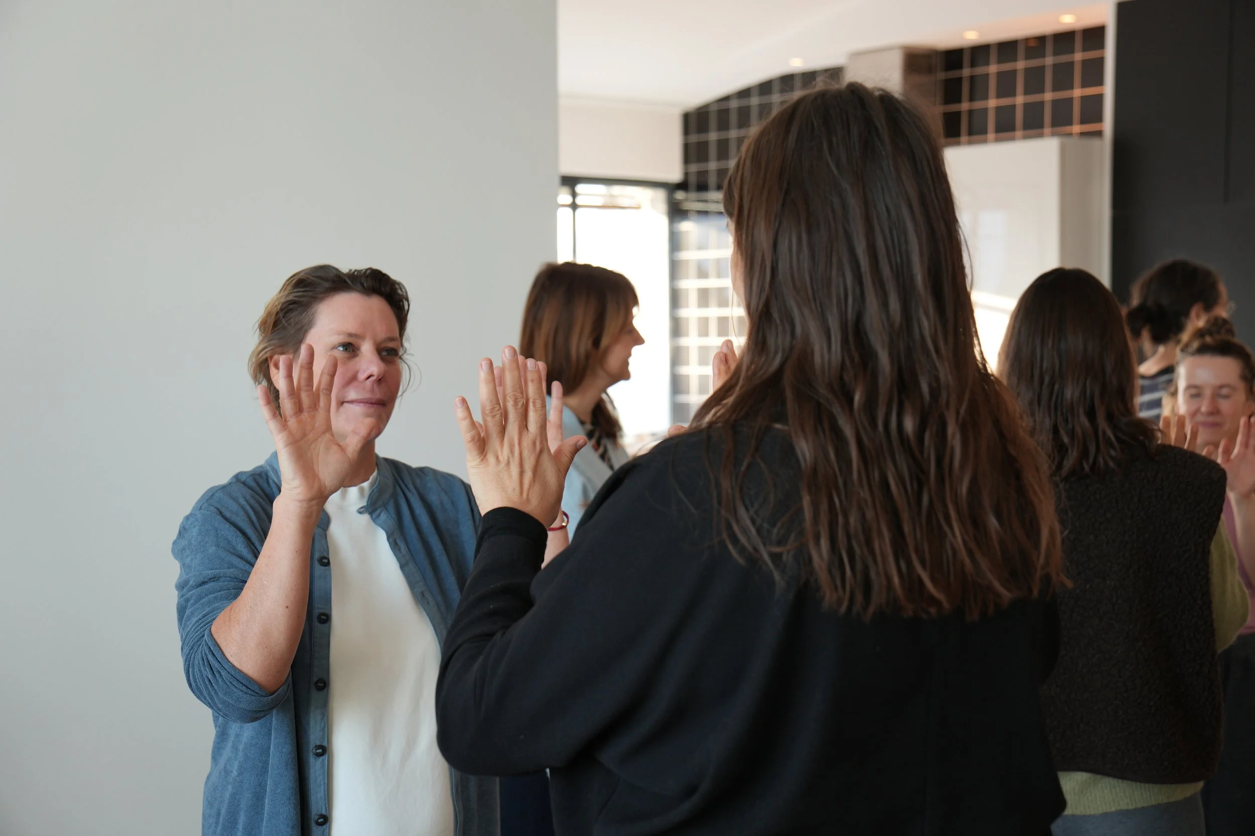 Zwei Frauen bei einem Händedruck und Blickkontakt in einem Raum mit mehreren Personen im Hintergrund.
