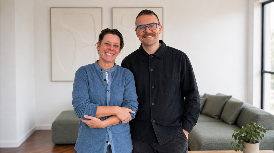 Ein Mann und eine Frau stehen nebeneinander in einem Wohnzimmer, beide lächeln. Im Hintergrund ist ein Sofa und ein Fenster.
