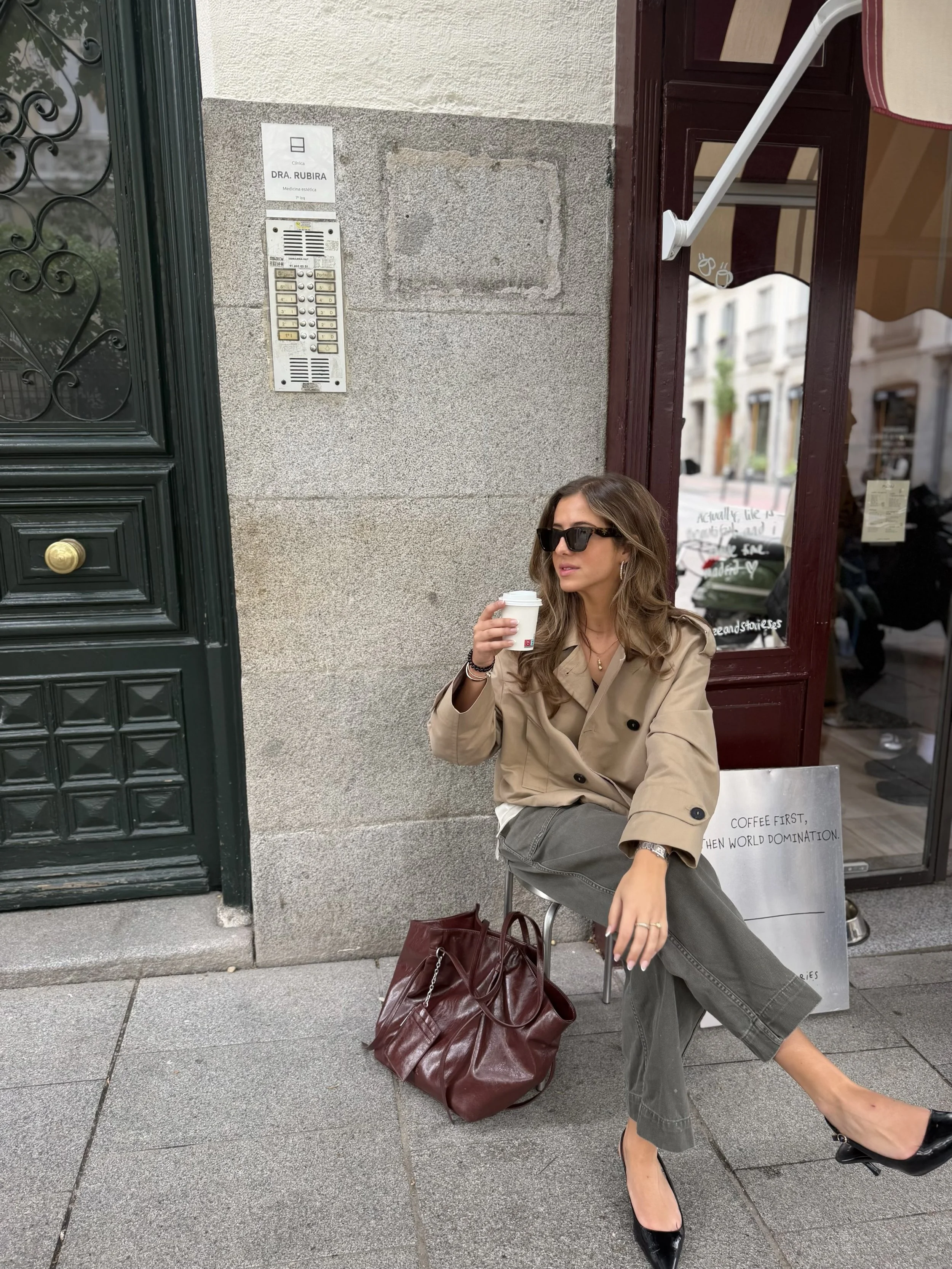 A woman with long wavy hair, wearing sunglasses, a beige trench coat, gray pants, and black high heels, sitting on a metal chair outside a shop, holding a coffee cup, with a large burgundy bag on the ground and a white shopping bag beside her that reads 'COFFEE FIRST, THEN WORLD DOMINATION.' She is sitting in front of a storefront with a glass door, and a person is reflected in the glass.