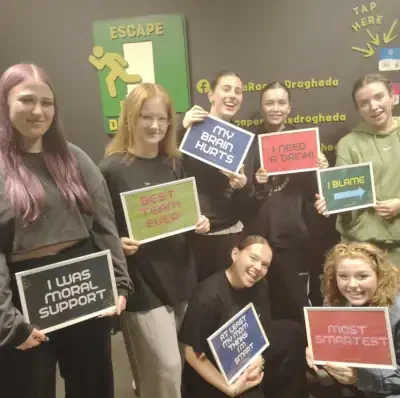 Seven young women at Escape Rooms Drogheda, holding signs with humorous and supportive messages, smiling and posing for a group photo.