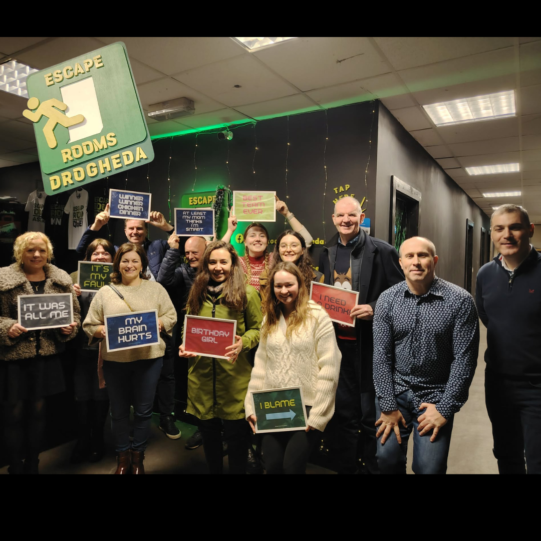 Group of people smiling and holding signs inside escape room called 'Drogheda' with exit sign and decorative lights in the background.