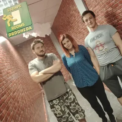 Three people standing in a brick-walled hallway, posing for a photo. A sign on the ceiling reads 'Escape Rooms Drogheda'.