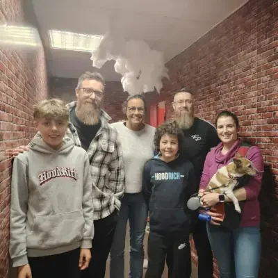 Group of six people standing together in a hallway with exposed brick walls and smoke or vapor coming from the ceiling, one person holding a small dog at Escape Rooms Drogheda