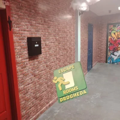 A corridor with brick walls, a red door on the left, a black mailbox or box mounted on the wall, and a sign that reads 'Escape Rooms Drogheda' on the floor. To the right, there are two doors and a colorful mural in the background.