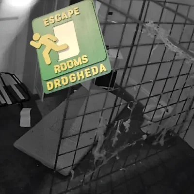 Escape Rooms Drogheda sign for rooms or a shelter, with a walking figure and the words 'ESCAPE ROOMS DROGHEDA', hanging on a metal fence. There is a spider and web on the fence.