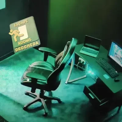 A computer desk setup with a black office chair, a laptop, a printer, and a desktop monitor, on a green carpet under dim lighting. A sign with a running figure and the words Escape Rooms Drogheda is on the side.