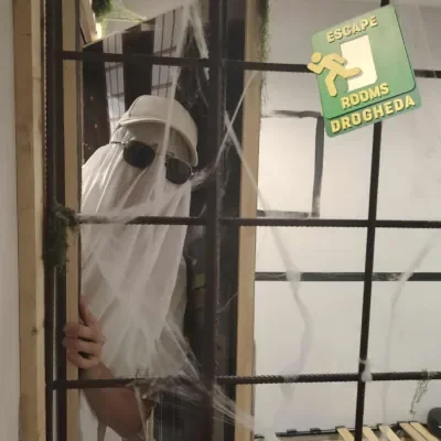 Person dressed as a ghost peeking through bars with a sign that reads "Escape Rooms Drogheda".