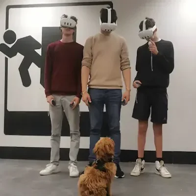 Three young men wearing virtual reality headsets standing in Escape Rooms Drogheda with a black and white wall design, with a brown dog sitting in front of them.