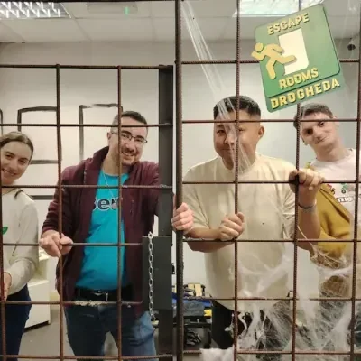 Four young people stand behind a metal cage with a sign that reads 'Escape Rooms Drogheda'.