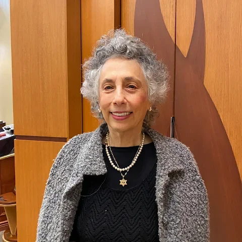 An elderly woman with curly gray hair, wearing a black top, a gray jacket, and pearl jewelry, standing indoors with wooden paneling in the background.