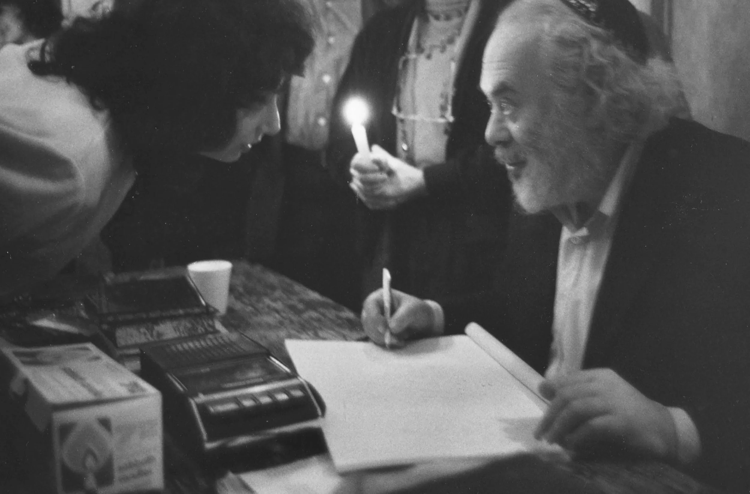 Two people, a woman and an older man, engage in a close, intimate interaction across a table with a calculator, papers, and a cup, in a black-and-white setting.