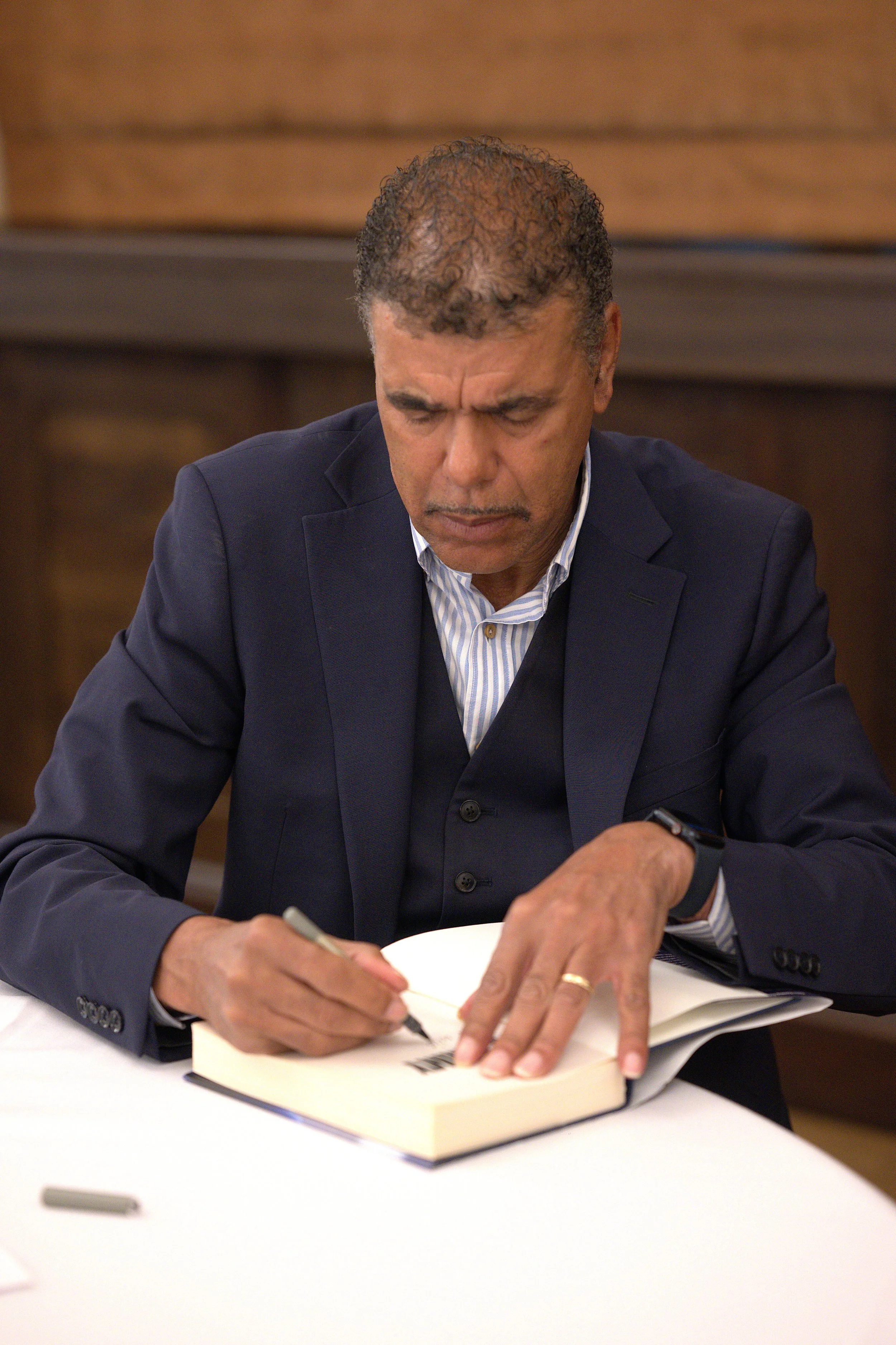 A man in a suit is sitting at a table, writing in a book, with a pen in his right hand and a smartphone on his left wrist. The background is a wall with wooden paneling.