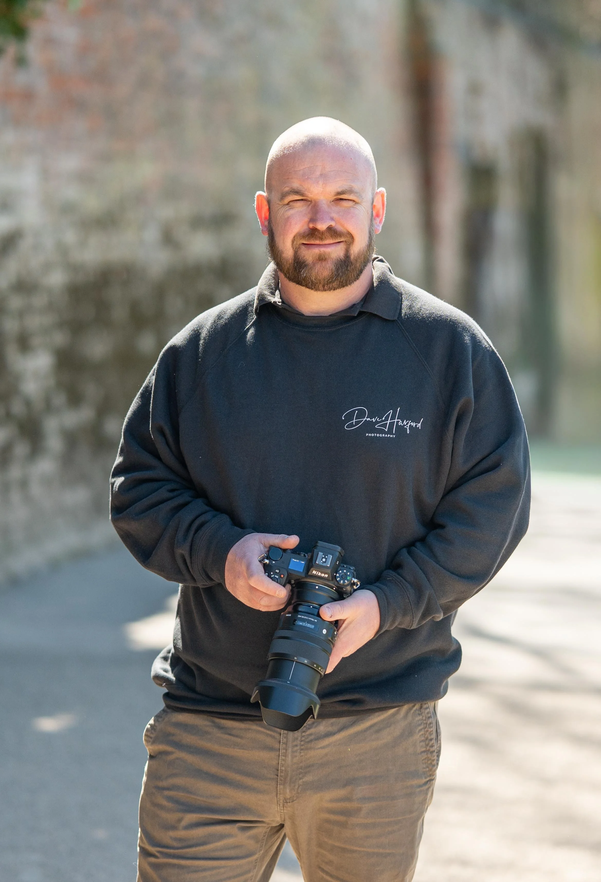 A man with a beard and no hair, wearing a black hoodie with the signature 'Dare Harford Photography' on it, holding a professional camera with a telephoto lens outdoors on a sunny day.