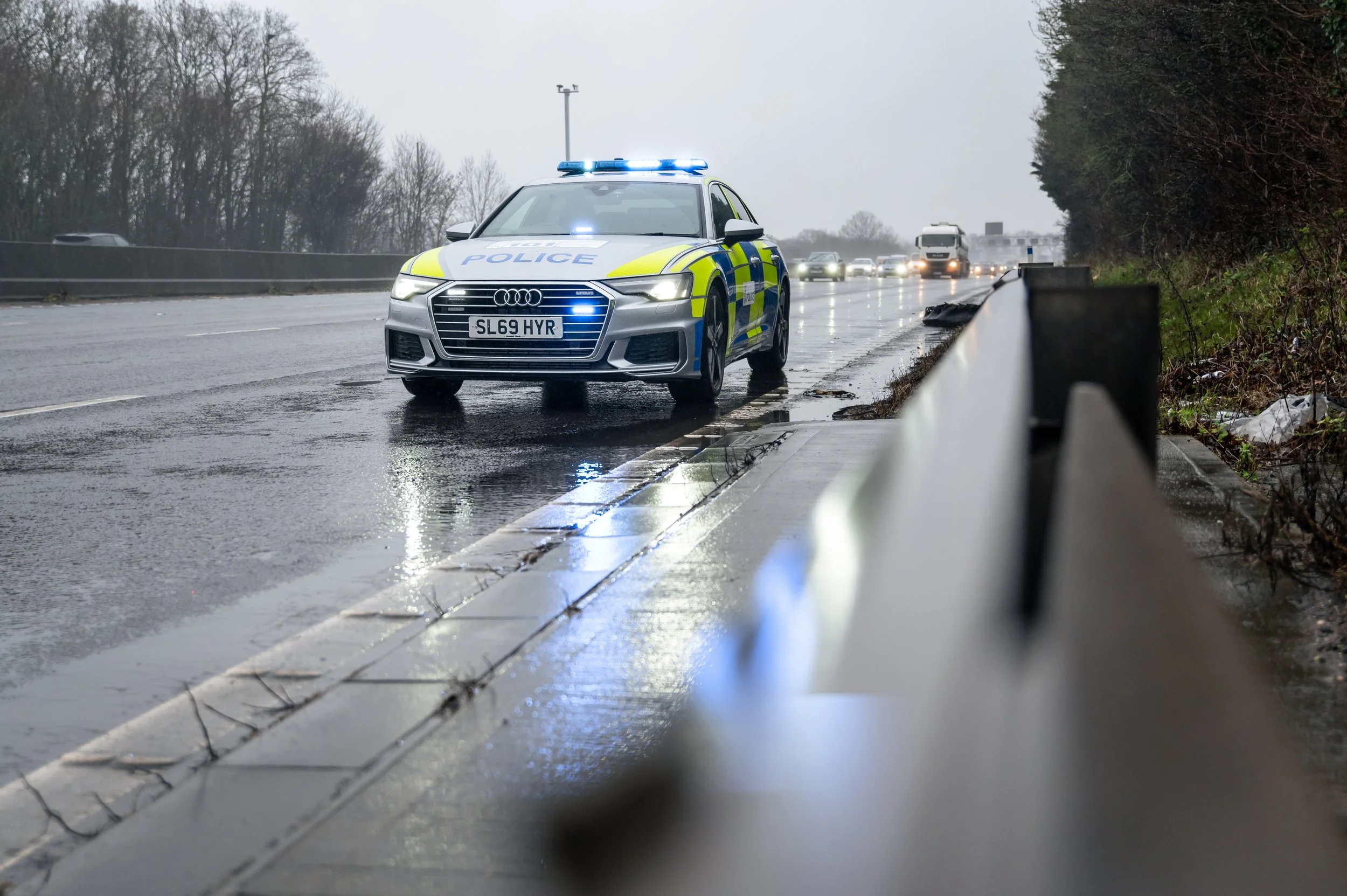 Police car on a wet road with blue emergency lights flashing, traffic and trees in the background, overcast weather.