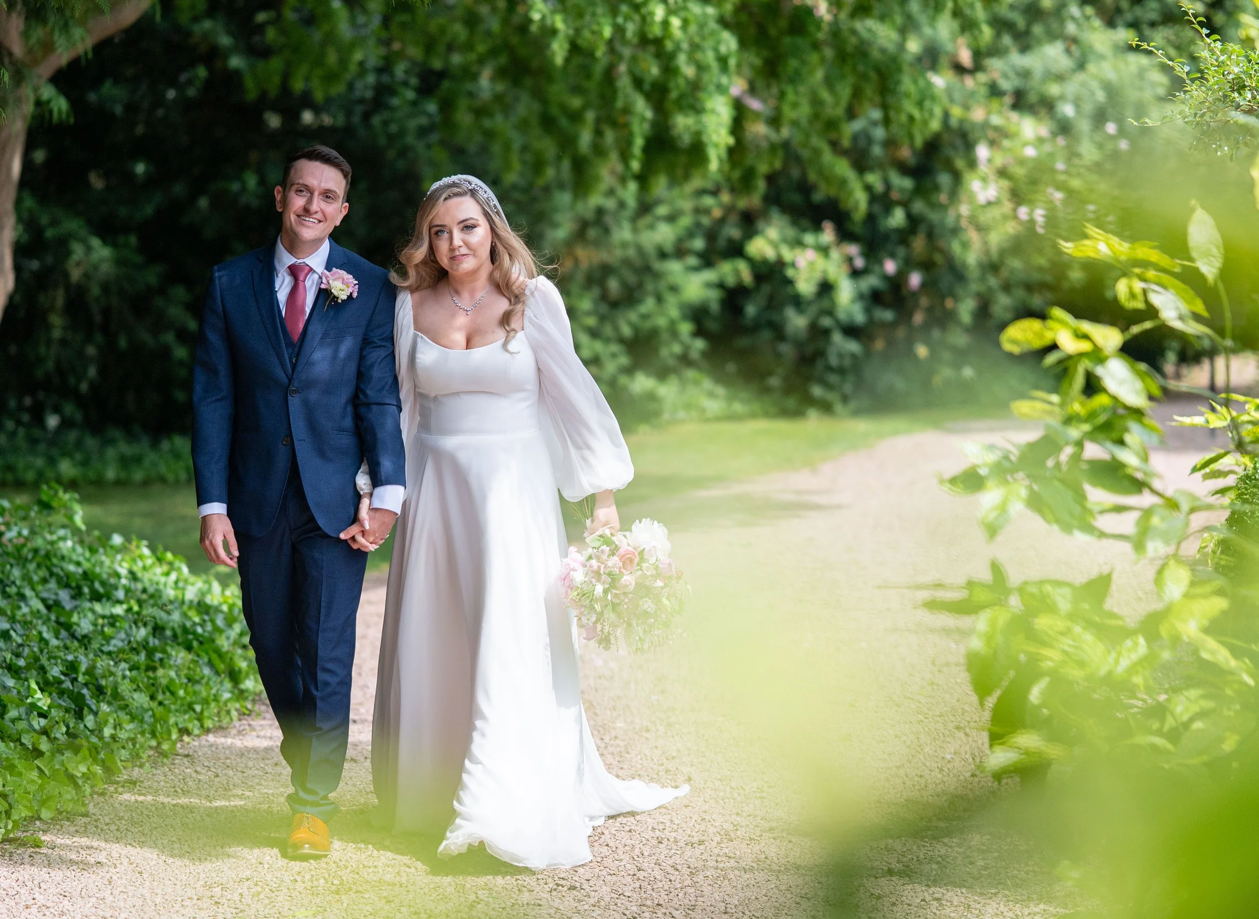 A newlywed couple walking arm in arm through a lush green park, smiling and looking at the camera. The groom is wearing a blue suit, red tie, and a boutonniere, while the bride is in a white wedding dress with a tiara and necklace, holding a bouquet of flowers.