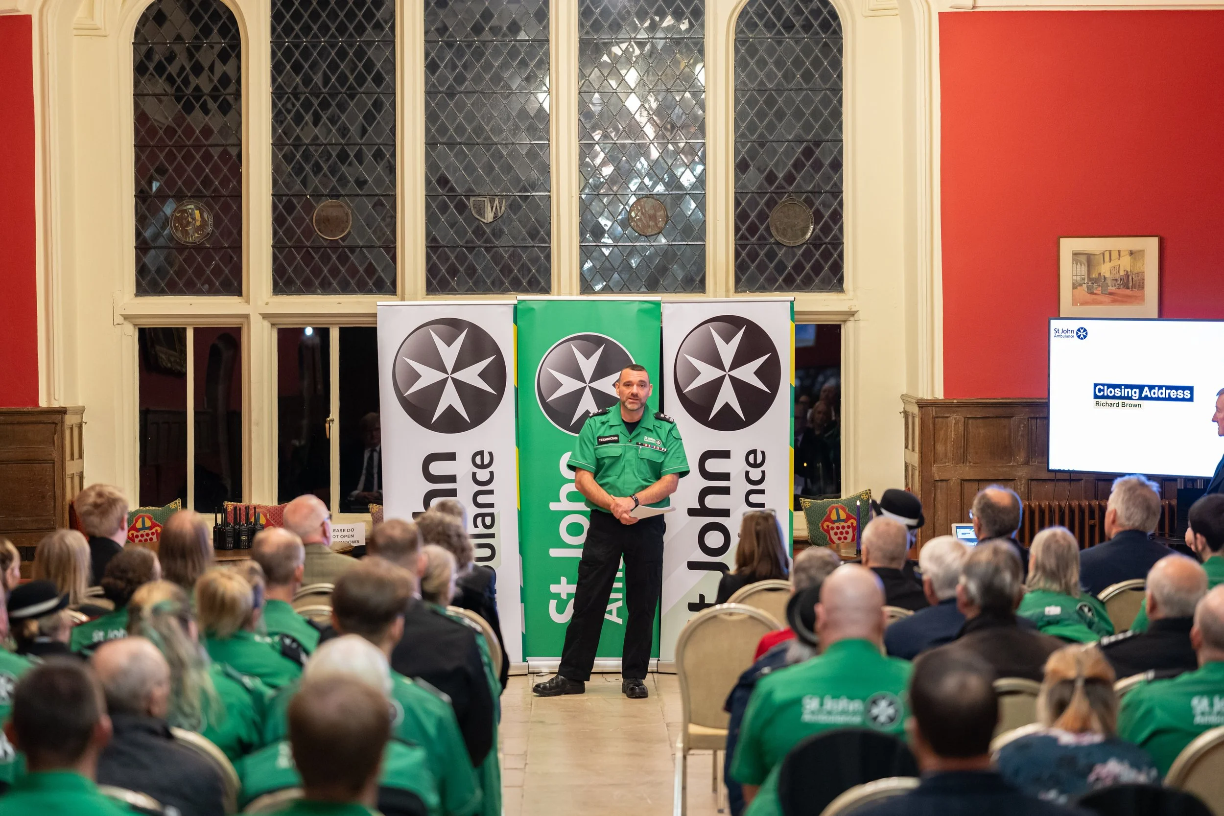 A man in a green uniform standing in front of a large group of people consisting mostly of witnesses wearing green uniforms, during a presentation or event at a historic building with stained glass windows. There are banners behind him with a black a