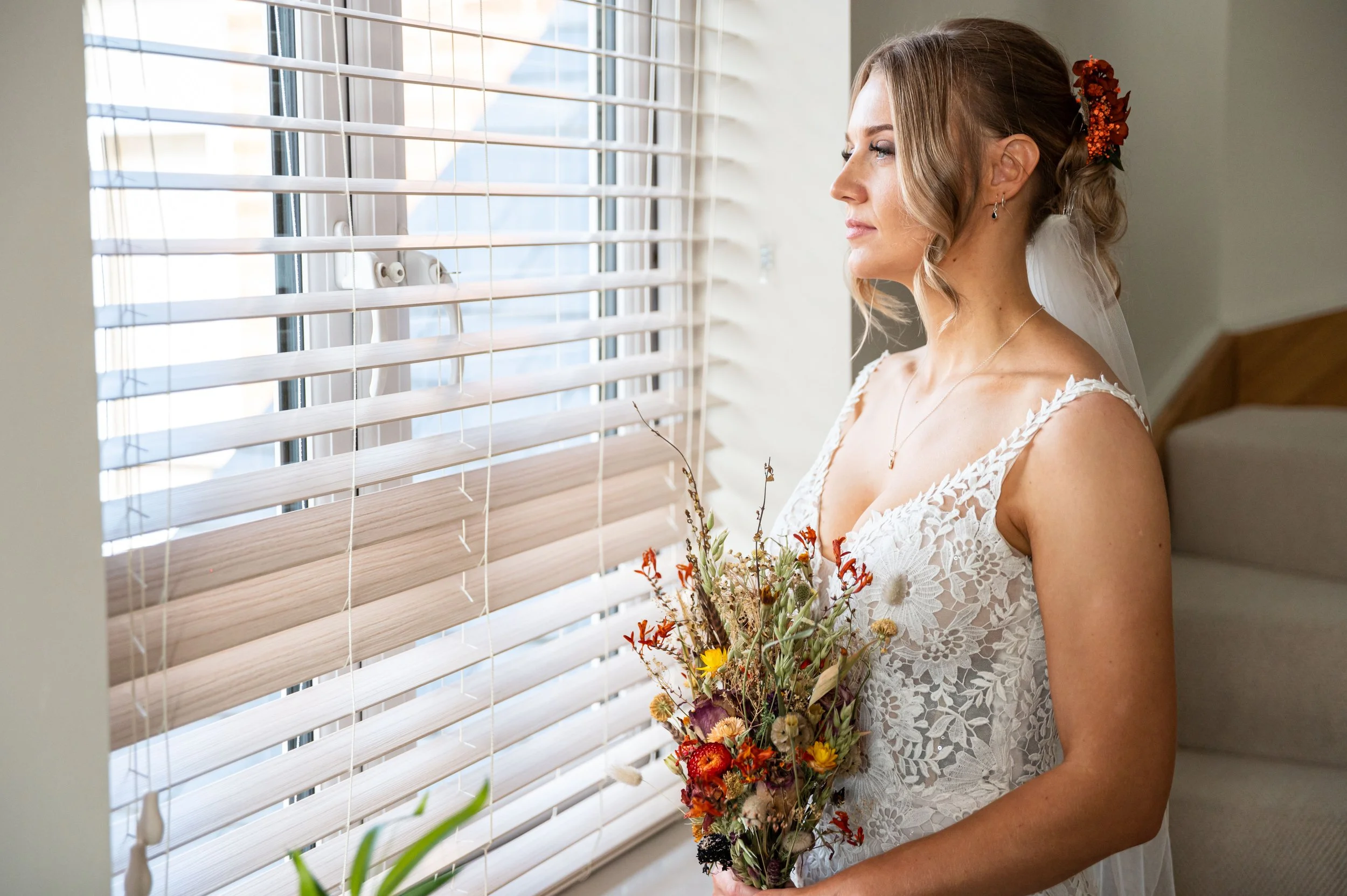 A bride standing indoors by a window with white blinds, holding a bouquet of colorful flowers, wearing a lace wedding dress, and looking out the window.