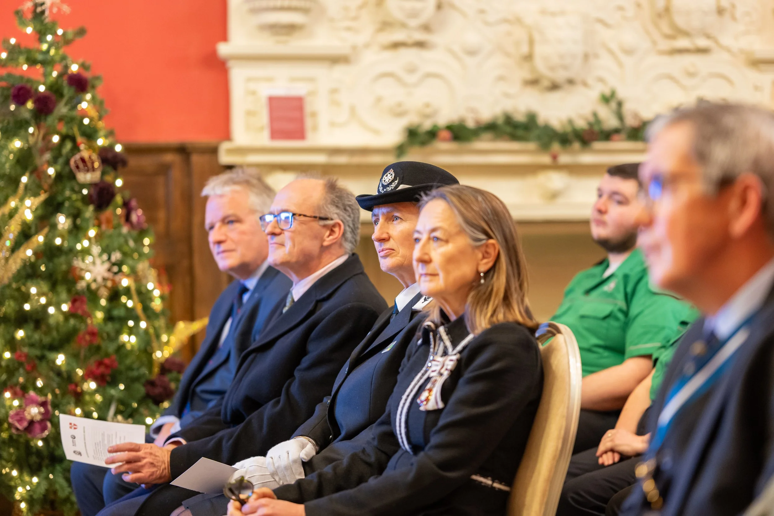People seated in chairs during a holiday event, with a decorated Christmas tree visible on the left side.