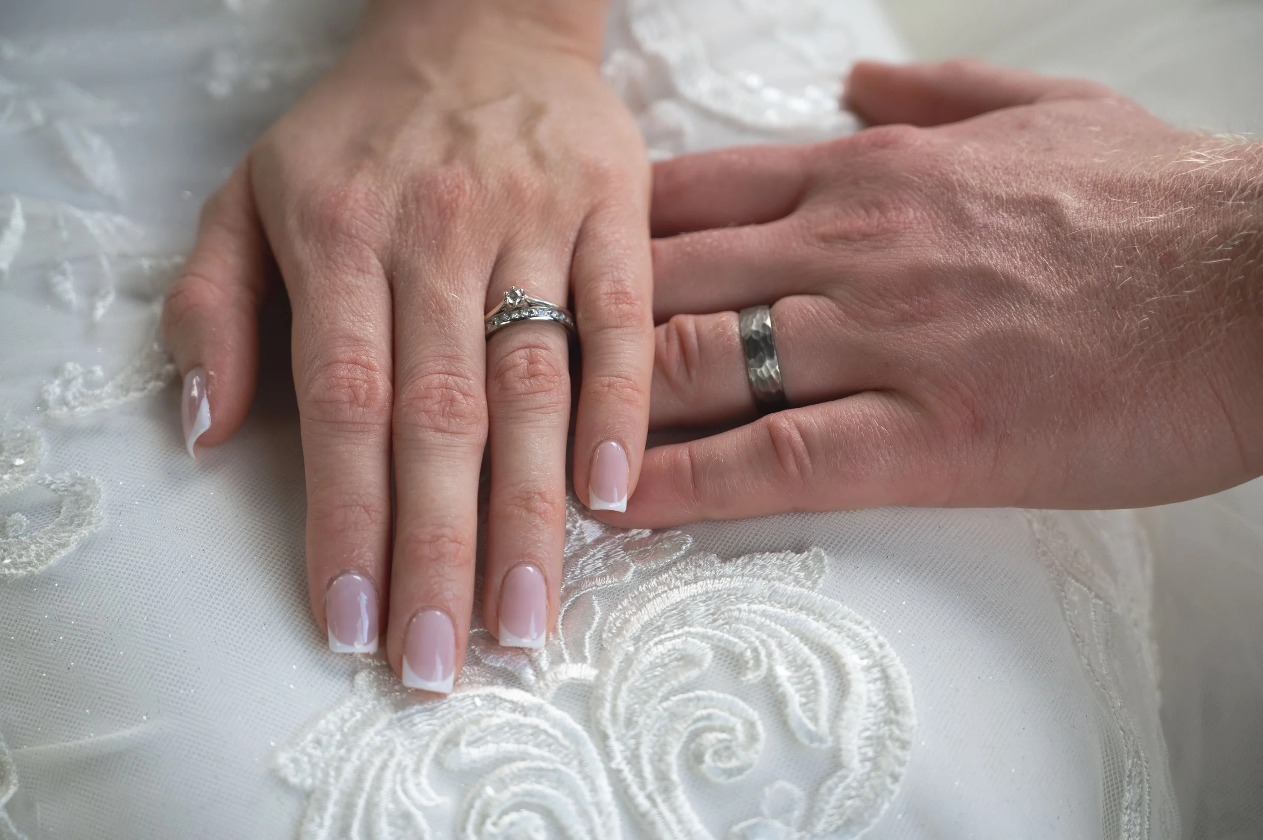 Close-up of a bride and groom's hands placed together on a white embroidered fabric, showing their wedding rings. The bride's hand has a diamond engagement ring and an anniversary band, while the groom's hand has a hammered silver wedding band.