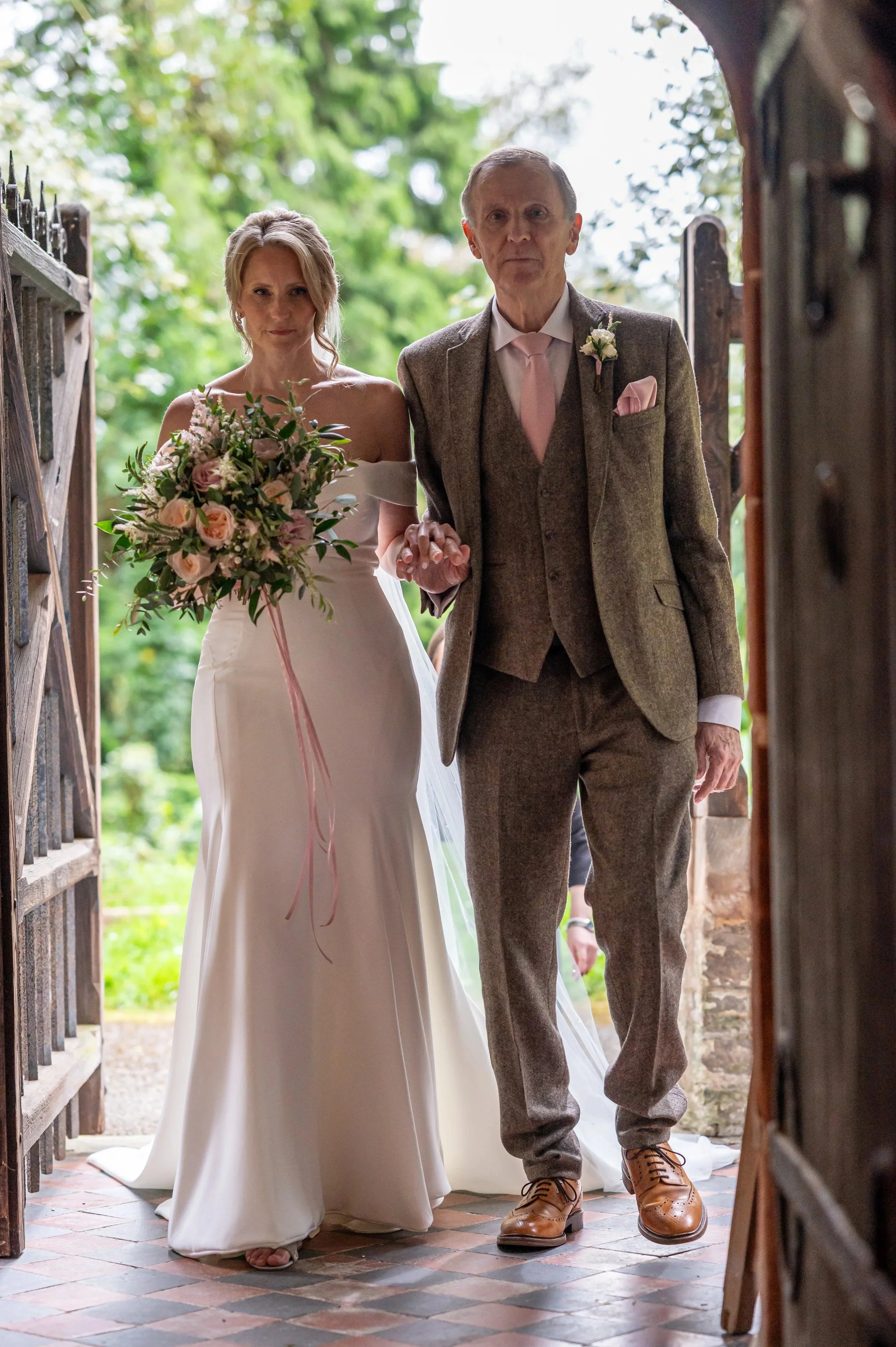 A bride and an older man, likely her father, walking out of a house or building on her wedding day. The bride is holding a bouquet of pink and white flowers, wearing a white wedding gown, and the man is dressed in a brown checkered suit with a pink tie and matching pocket square. They are holding hands.