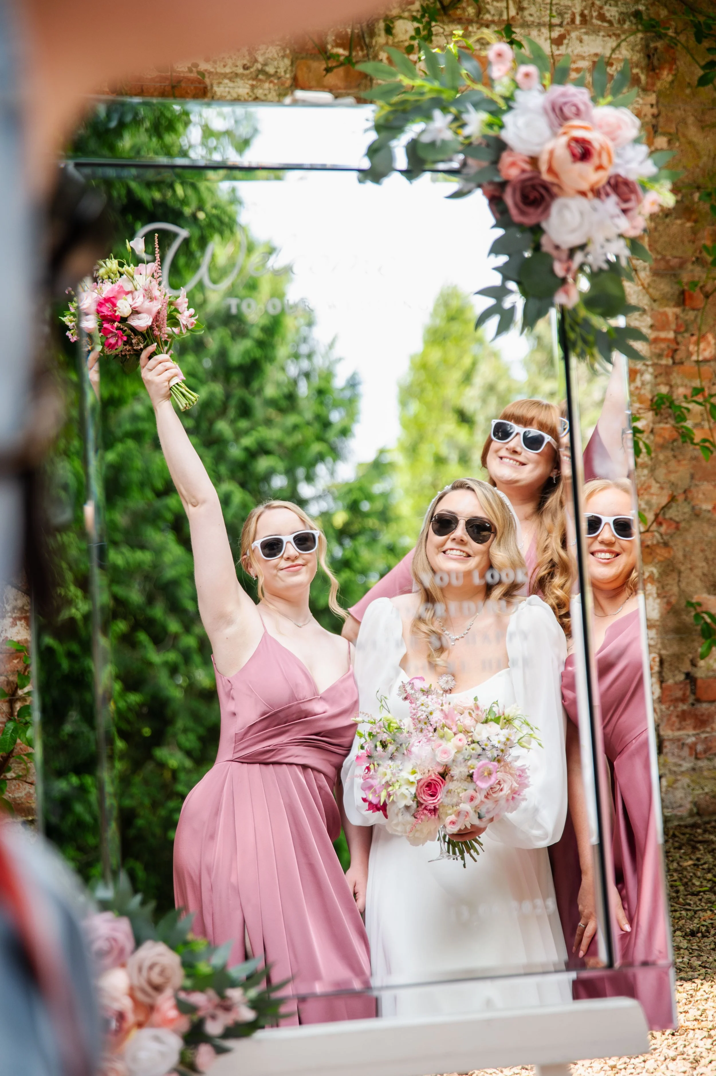 A group of women, including a bride in a white dress holding a bouquet of pink and white flowers, taking a selfie in front of a mirror outdoors. They are wearing sunglasses, and some are dressed in pink dresses. The scene is decorated with floral arrangements and greenery on a brick wall.