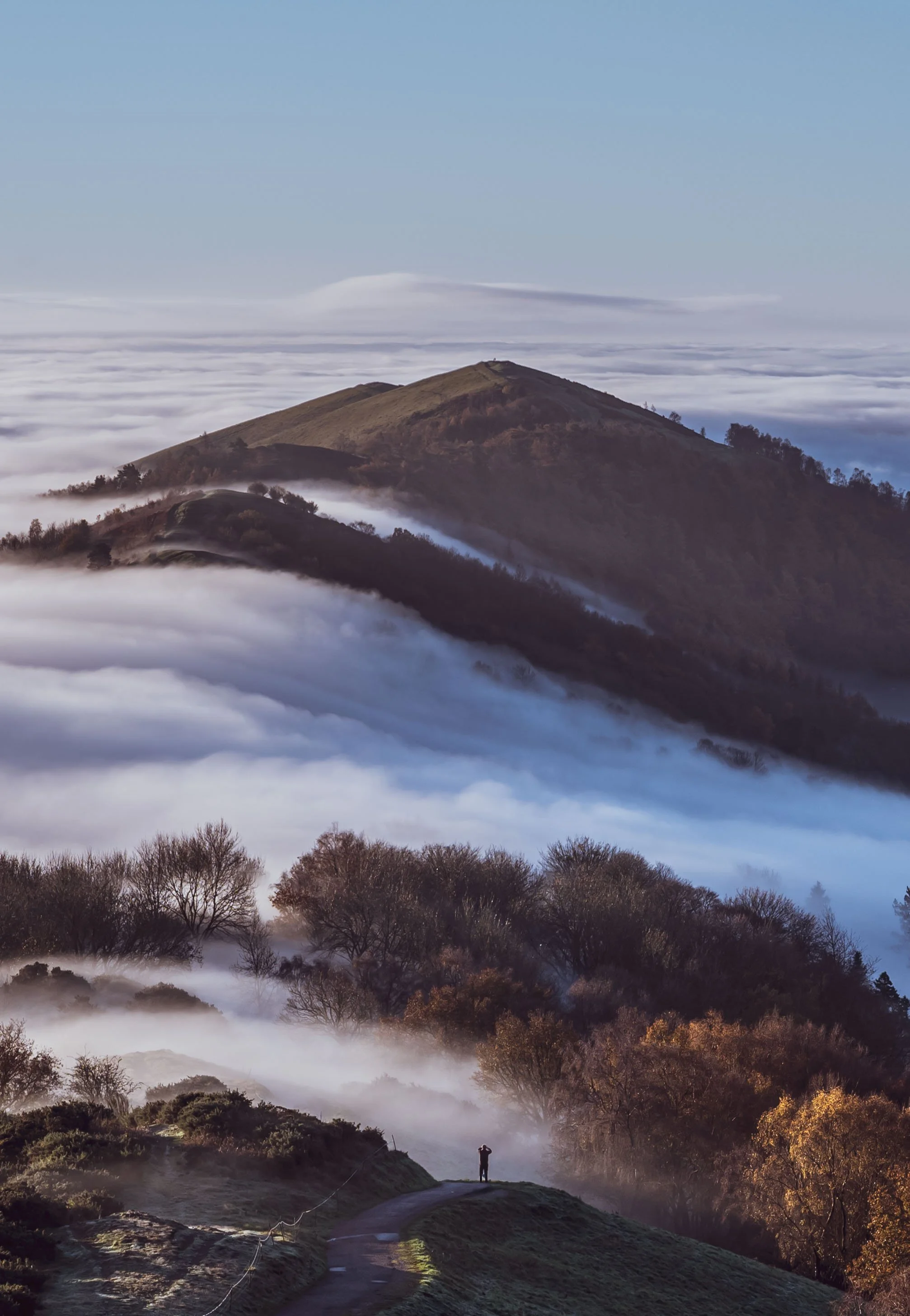 A person standing on a path on a mountain surrounded by mist and clouds, with hills and trees in the background under a blue sky.