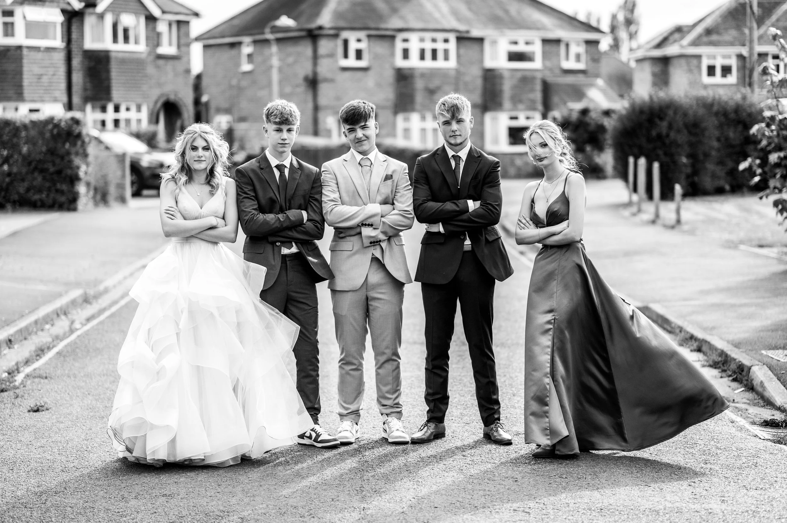 Black and white photo of five young adults standing on a street, dressed in formal attire, with a row of houses in the background.