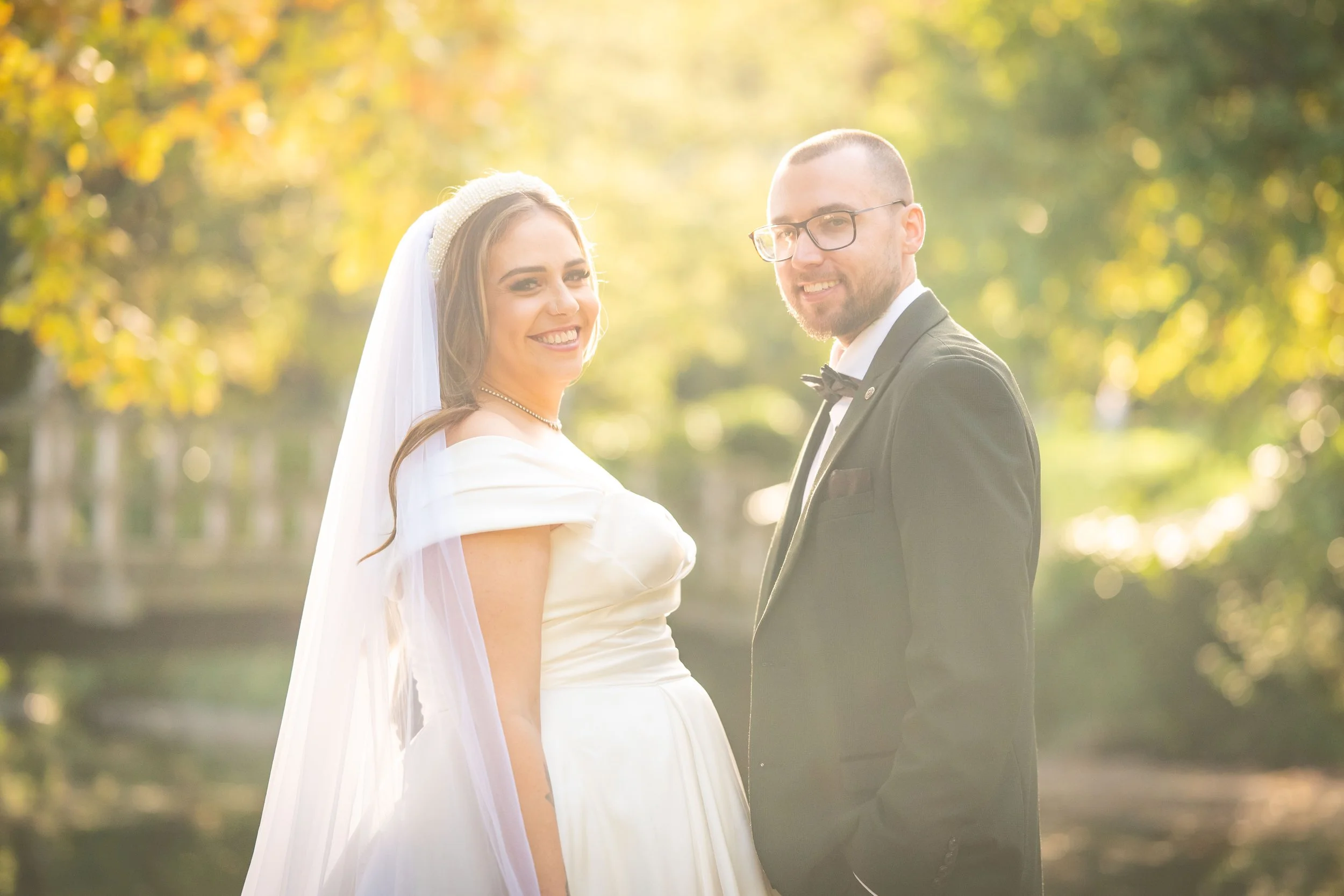 A bride and groom standing outdoors surrounded by trees during daytime, smiling at the camera. The bride wears a white wedding dress and veil, and the groom wears a black tuxedo with a bow tie and glasses.